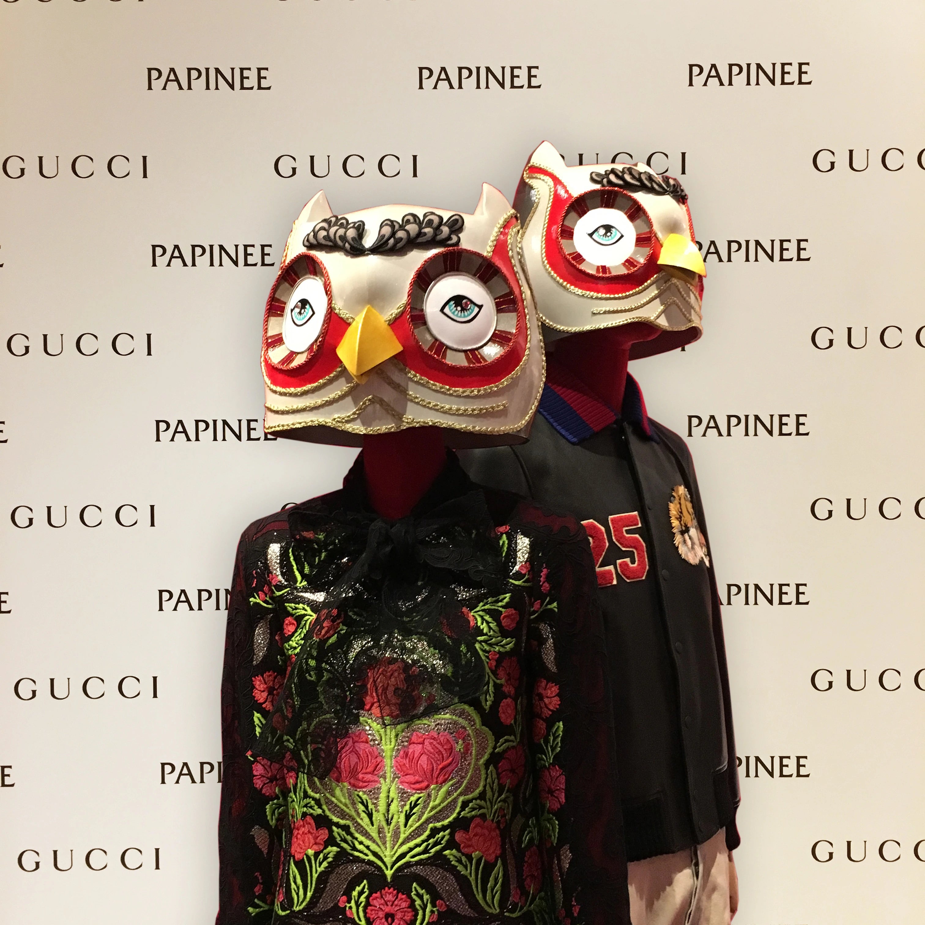 Two people wearing large owl masks in front of a Gucci and Papinee backdrop.